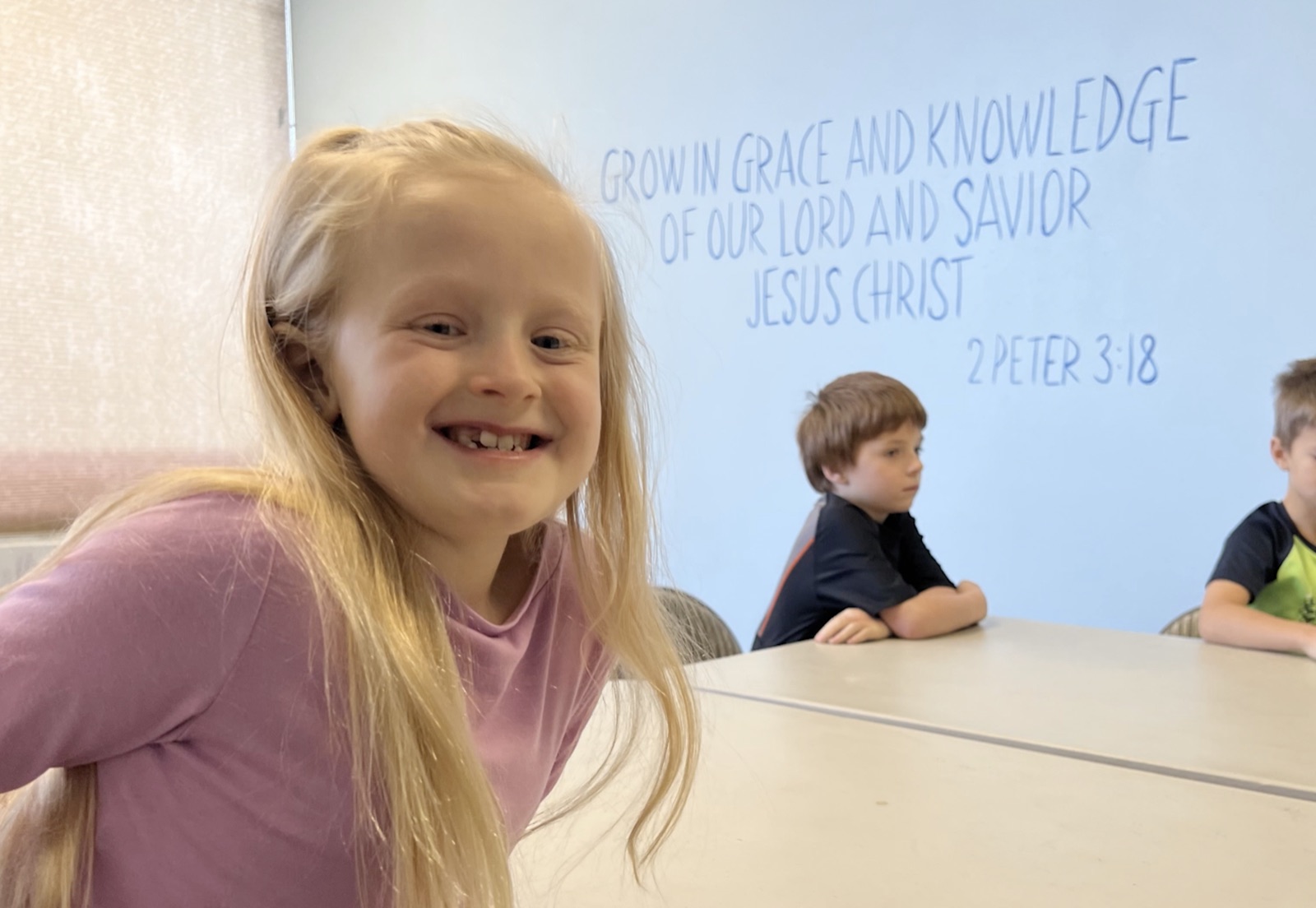 A girl smiling in the Grace Kids room, in front of a wall painted with 2 Peter 3:18