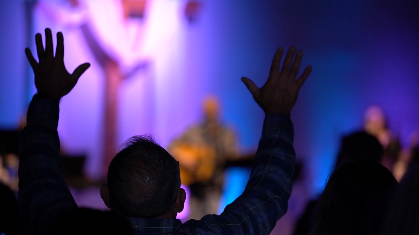 A man with both hands raised in worship during a Sunday service, lit in blue and purple